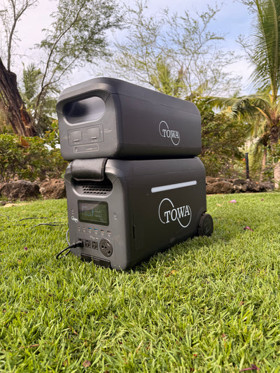 Two black TOWA portable power stations stacked outdoors on a green lawn with trees and shrubs in the background.